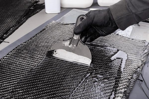 Gloved hand applying resin to a carbon fiber sheet with a plastic scraper.