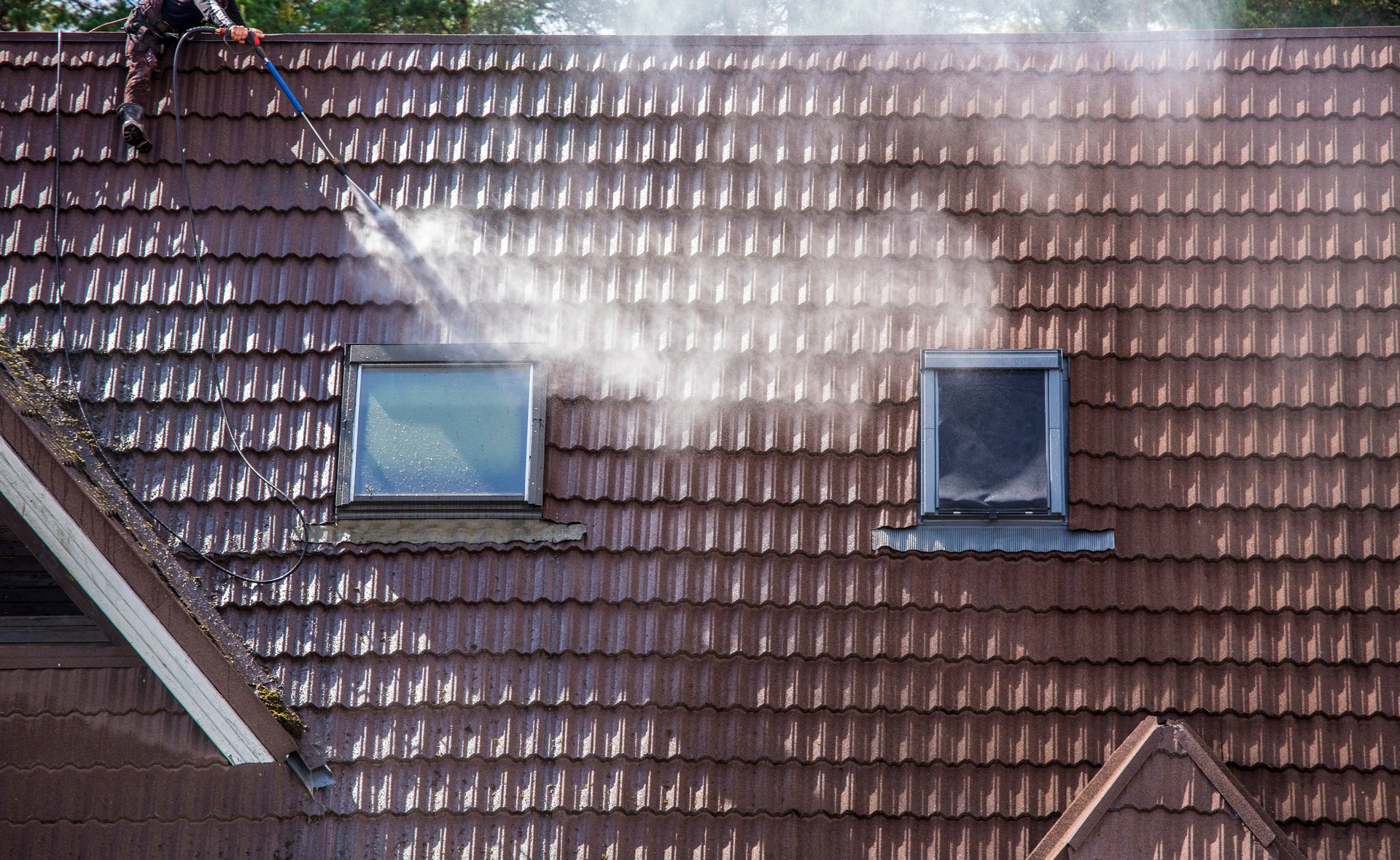 Man worker washing tilted stone roof shingles with water pressure washer to remove dirt, moss and lichen outdoors in summer. Roof cleaning service concept.